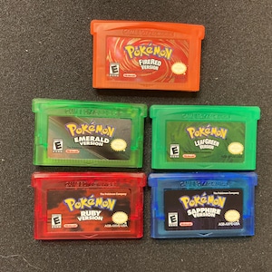 Könnte beinhalten: Fünf Game Boy Advance Videospielkassetten, darunter die Versionen Pokémon FireRed, Emerald, LeafGreen, Ruby und Sapphire. Die Kassetten sind orange, grün und blau und zeigen jeweils das Pokémon-Logo, die ESRB-Bewertung und das Nintendo-Logo.