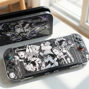 May include: A handheld gaming console and its docking station featuring a black and white manga-style collage print. The console displays various character illustrations and the text NANA and BLACK STONES. The dock features a purple and black floral design.