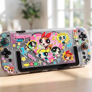 Pode incluir: Uma consola de jogos portátil com uma capa protetora transparente decorada com autocolantes das Powerpuff Girls. O design inclui um grande coração cor-de-rosa, várias ilustrações de personagens, estrelas coloridas, relâmpagos e o texto The Powerpuff Girls.
