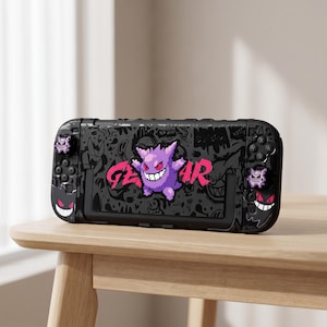 Op de afbeelding: Een zwarte beschermhoes voor een draagbare spelcomputer met een gepixelde paarse Gengar-figuur in het midden en de tekst GENGAR in felroze. De hoes bevat bijpassende paarse Gengar-joystickdoppen en kleine Gengar-afbeeldingen op de zijkanten.