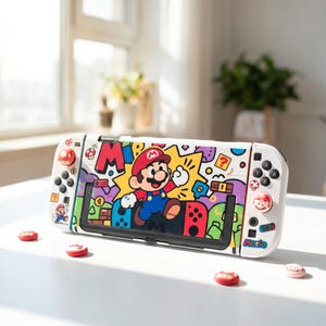 Op de afbeelding: Een witte draagbare videogameconsole met een kleurrijke afbeelding van Mario op het middenpaneel. Het apparaat bevat witte controllers met rood-witte paddenstoelvormige joystickdoppen, zwarte knoppen en extra Mario-stickers.