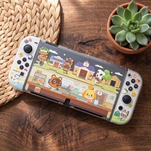 May include: A handheld gaming console featuring a clear protective case decorated with Animal Crossing characters including Tom Nook, Isabelle, and K.K. Slider. The white controllers are adorned with small colorful icons like cherries, stars, and leaves.