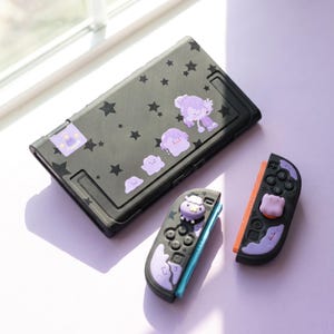 Könnte beinhalten: Eine schwarze Spielkonsole und zwei Controller mit violetten Sternenmustern und hellvioletten Charakteraufklebern. Die Konsole ist rechteckig, während die Controller hellviolette Stick-Kappen und dekorative Akzente aufweisen.