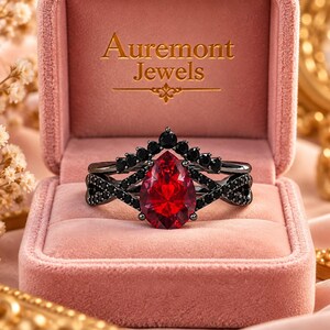 May include: A black metal ring set featuring a pear-shaped red center stone and small black accent stones. The ring is displayed in a pink velvet box with the text Auremont Jewels printed in gold on the inside of the lid.
