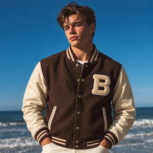 Puede incluir: Una chaqueta universitaria marrón con mangas de color crema, un parche grande con la letra B de color crema en el pecho izquierdo, cuello, puños y dobladillo acanalados con rayas crema, y cierre frontal con botones metálicos sobre una camisa blanca.