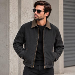 Puede incluir: Una chaqueta de lona gris carbón con cuello de pana marrón en contraste, cremallera frontal de metal y dos bolsillos laterales. La chaqueta se lleva sobre una camiseta negra de cuello redondo, combinada con vaqueros oscuros y gafas de sol.