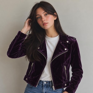 May include: A deep purple velvet biker jacket featuring a notched collar, silver-toned metal zips on the front and pockets, and silver snap button accents on the lapels and hem, worn over a plain white crew neck t-shirt and blue denim jeans.