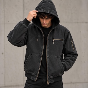 May include: A charcoal grey denim hooded jacket featuring a full-length front zipper, a drawstring hood, ribbed cuffs and hem, two lower front pockets, a horizontal zippered chest pocket, and an additional zippered utility pocket on the left sleeve.