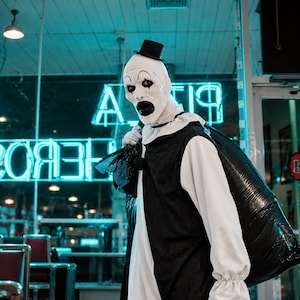 Peut inclure: Un costume de clown composé d'une chemise blanche à manches longues avec des poignets à volants, d'un gilet noir et d'un haut-de-forme noir. Le clown a le visage blanc avec du maquillage noir aux yeux et à la bouche, portant un grand sac poubelle noir.