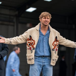 Könnte beinhalten: Eine cremefarbene, grob gestrickte Strickjacke mit Reißverschluss und einem auffälligen Fuchsmotiv auf den Vorderteilen, getragen über einem hellblauen Hemd und einer hellblauen Jeans. Die Strickjacke hat einen Schalkragen, Rippbündchen und zwei Taschen.