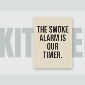 May include: A rectangular wall sign with a light beige background and bold black text that reads: THE SMOKE ALARM IS OUR TIMER. The sign is positioned over a light gray background featuring the partially visible, large gray letters K I T.