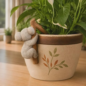 May include: A beige ceramic planter featuring a textured koala figure clinging to a brown branch attached to the side. The planter has a dark brown rim and a painted green and orange leaf design on the front, containing a green leafy plant.
