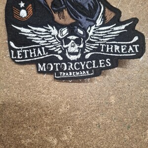 Pu&ograve; includere: Una toppa ricamata nera raffigurante un teschio con occhiali protettivi e ali, il testo LETHAL THREAT MOTORCYCLES TRADEMARK e un'insegna a gallone in stile militare con una stella, realizzata con filo bianco, giallo e marrone su fondo nero.