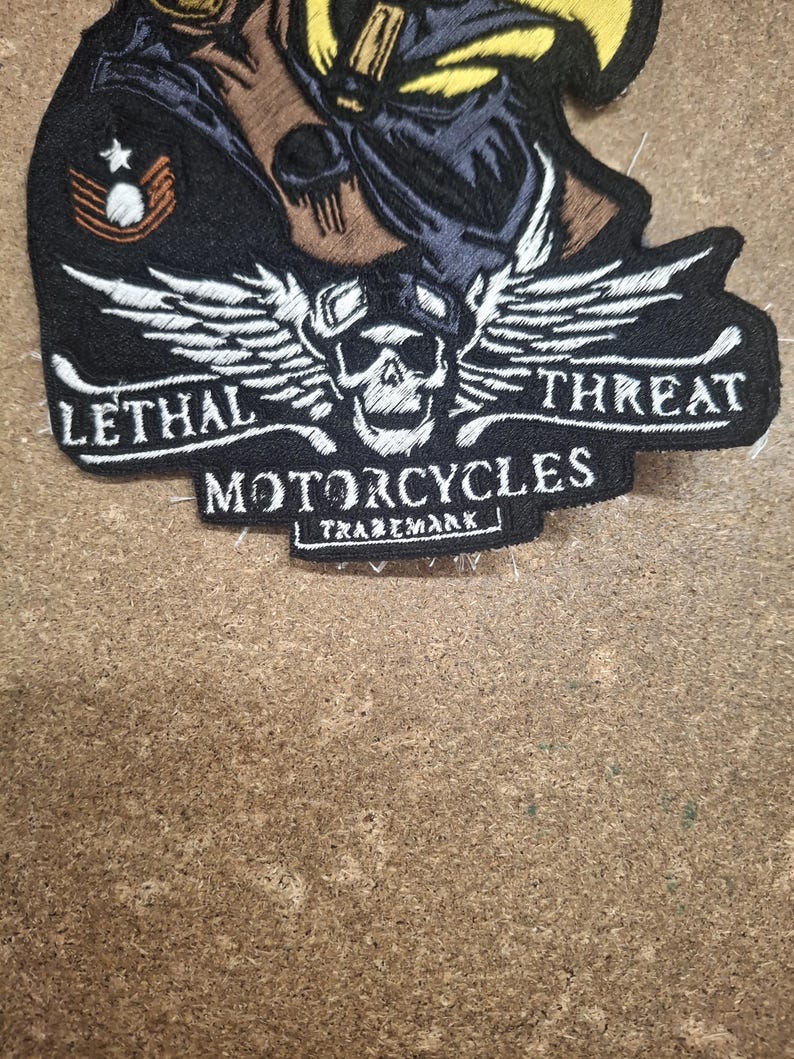 Pu&ograve; includere: Una toppa ricamata nera raffigurante un teschio con occhiali protettivi e ali, il testo LETHAL THREAT MOTORCYCLES TRADEMARK e un'insegna a gallone in stile militare con una stella, realizzata con filo bianco, giallo e marrone su fondo nero.