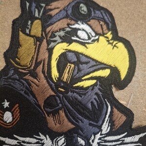 Pu&ograve; includere: Una toppa ricamata raffigurante una testa d'aquila stilizzata che indossa un berretto da aviatore marrone e occhiali protettivi. L'aquila ha un becco giallo brillante e piumaggio scuro. Sotto l'aquila c'&egrave; un teschio bianco con ali ed emblema.