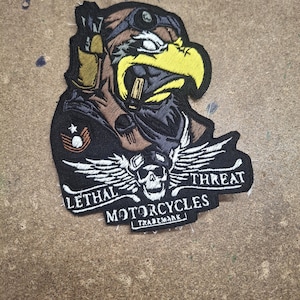 Può includere: Una toppa ricamata raffigurante un'aquila stilizzata con occhiali protettivi e casco, con un emblema a forma di teschio e ali sottostanti. Il testo recita LETHAL THREAT MOTORCYCLES TRADEMARK in filo bianco su sfondo nero con accenti gialli e marroni.