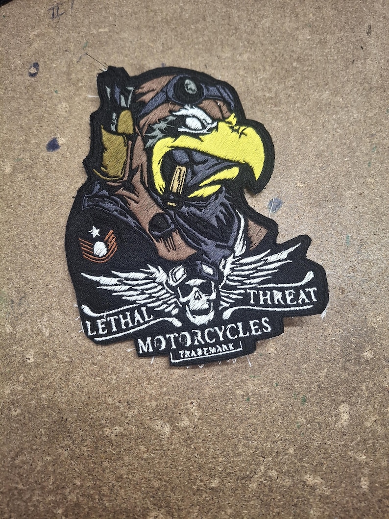 Pu&ograve; includere: Una toppa ricamata raffigurante un'aquila stilizzata con occhiali protettivi e casco, con un emblema a forma di teschio e ali sottostanti. Il testo recita LETHAL THREAT MOTORCYCLES TRADEMARK in filo bianco su sfondo nero con accenti gialli e marroni.
