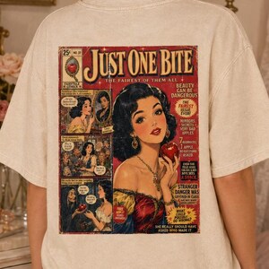 May include: A beige short-sleeve t-shirt featuring a large graphic on the back designed like a vintage comic book cover titled Just One Bite. The illustration depicts a woman with dark hair holding a red apple, surrounded by comic panels.