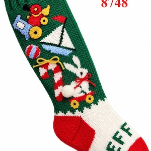 Può includere: Una calza natalizia in maglia verde con la scritta TOYLAND 8748. La calza è decorata con un trenino, una barca a vela, una palla a righe, un bastoncino di zucchero e un coniglio bianco su uno skateboard. Il piede è bianco con il nome JEFF in verde.