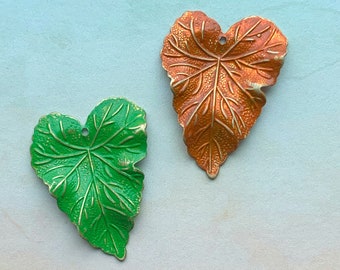 May include: Two heart-shaped leaf pendants featuring embossed vein details. One pendant is finished in a vibrant green enamel with metallic gold highlights, while the other is finished in a warm copper-orange enamel with similar gold-toned accents.