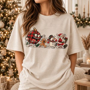May include: A heathered beige t-shirt featuring a graphic print of a red and green plaid Christmas ornament with an angry face chasing a gingerbread man, a snowman, and Santa Claus, who is holding a wrapped gift while running away.