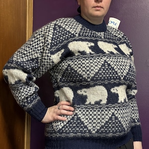 Peut inclure: Un pull à manches longues et col rond arborant un motif répétitif d'ours polaires blancs sur fond bleu marine foncé. Le tricot présente des motifs géométriques triangulaires ainsi qu'un col, des poignets et un ourlet côtelés bleu marine.