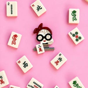 May include: A brooch shaped like a person with a red hair bow, black glasses, and red lips, holding a mahjong tile with the characters 二萬. The brooch is surrounded by various scattered mahjong tiles on a solid pink background.
