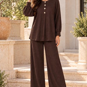 May include: A dark brown two-piece set consisting of a long-sleeved top and wide-leg trousers. The top features a round neckline, a partial button-down front with four light-coloured buttons, and gathered cuffs. The fabric has a vertical ribbed texture.