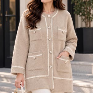 May include: A beige long-sleeved cardigan with a fuzzy texture, featuring a round neckline, a front button closure, and four patch pockets. The garment is accented with white trim along the placket, pockets, and cuffs, each finished with a small button.