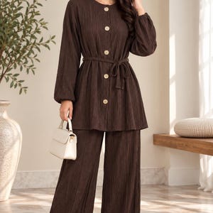 May include: A dark brown two-piece set consisting of a long-sleeved tunic with a round neckline, five light-coloured buttons down the front, and a matching tie belt, paired with wide-leg trousers. A small cream-coloured handbag is held in one hand.