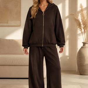 May include: A dark brown matching set consisting of a long-sleeve zip-up hoodie with a gold-toned circular zipper pull and wide-leg trousers. The hoodie features a hood, ribbed cuffs, and a hem, while the trousers have a relaxed, loose fit.