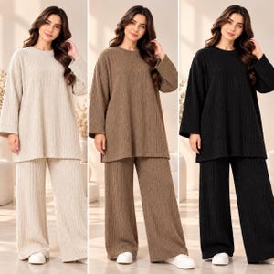 May include: A two-piece lounge set consisting of a long-sleeved, round-neck tunic and wide-leg trousers. The garments feature a vertical ribbed texture and are presented in three distinct solid colours: beige, brown, and black.