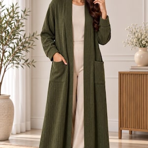 Könnte beinhalten: Eine lange, offene Strickjacke in Olivgrün aus strukturiertem Rippenstoff mit langen Ärmeln, Bündchen und zwei großen aufgesetzten Taschen auf der Vorderseite. Das Kleidungsstück wird über einem beigen Oberteil, einer beigen Hose und weißen Sneakern getragen.