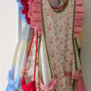 May include: A collection of aprons hanging on hangers, featuring various floral and patterned fabrics with ruffled shoulder straps and large fabric bows at the waist. The aprons include designs in pink floral, red, and light blue solid colours.