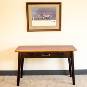 May include: A rectangular wooden desk with a natural light brown top and a dark brown base featuring a single central drawer with a thin horizontal gold-toned metal handle. Above the desk hangs a framed landscape print of a snowy farm scene.