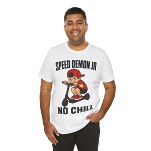 Puede incluir: Una camiseta blanca de manga corta con un gráfico de un personaje de dibujos animados con gorra y camiseta rojas montando un patinete negro. El texto SPEED DEMON JR está impreso en negro arriba y el texto NO CHILL está impreso en negro abajo.