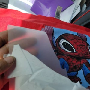 Puede incluir: Una camiseta roja con un personaje de dibujos animados azul y rojo que lleva un disfraz de Spiderman. El personaje tiene ojos grandes y una gran sonrisa. Una l&aacute;mina de pl&aacute;stico transparente se est&aacute; despegando de la camiseta.