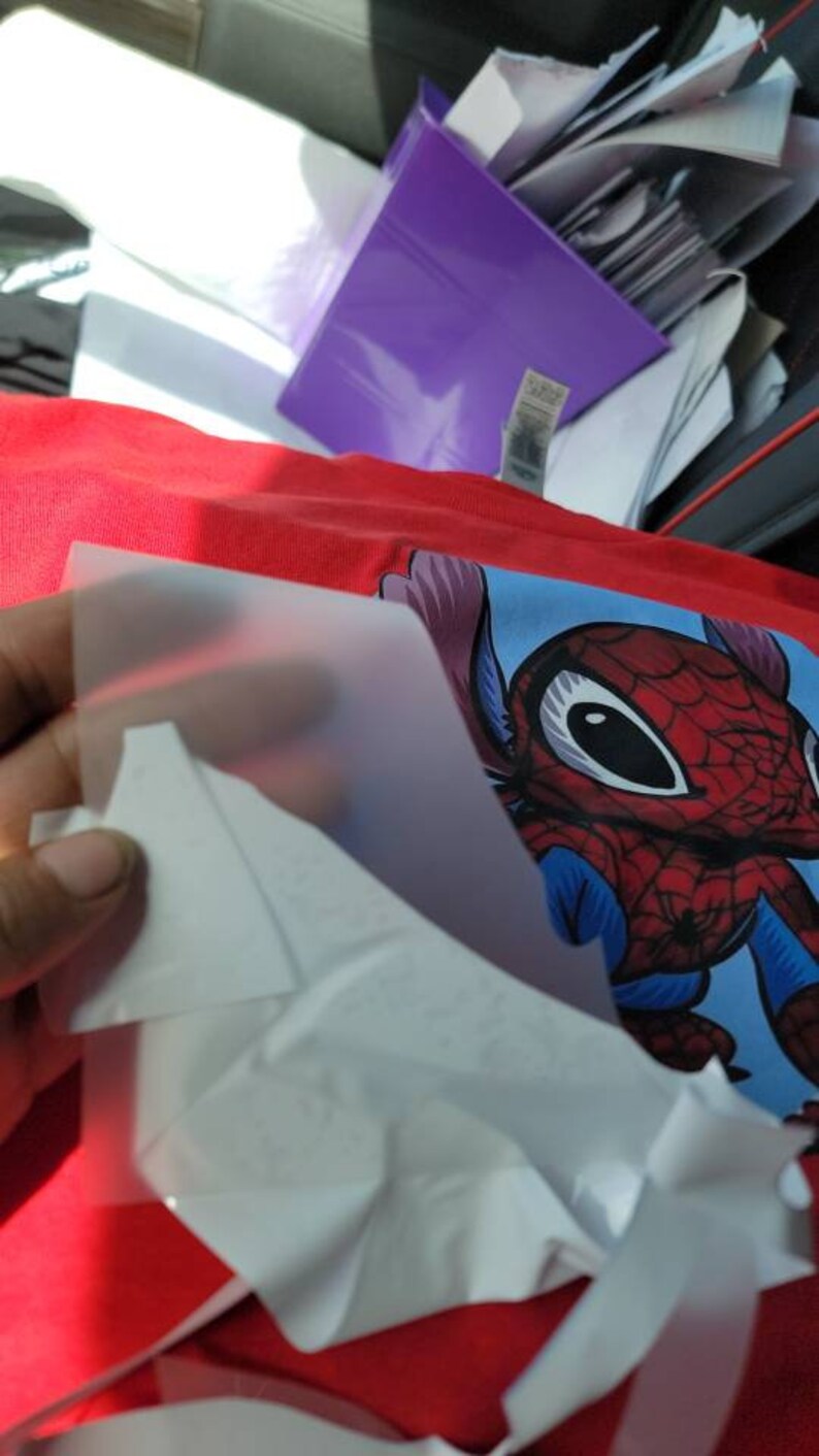 Puede incluir: Una camiseta roja con un personaje de dibujos animados azul y rojo que lleva un disfraz de Spiderman. El personaje tiene ojos grandes y una gran sonrisa. Una l&aacute;mina de pl&aacute;stico transparente se est&aacute; despegando de la camiseta.