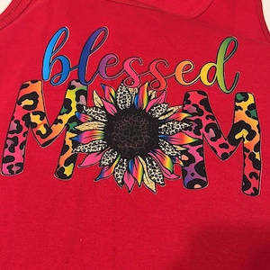 Puede incluir: Camiseta de tirantes roja con las palabras "Blessed Mom" en colores arco&iacute;ris con un girasol en el medio. El girasol tiene un centro negro y p&eacute;talos arco&iacute;ris con un estampado de leopardo.