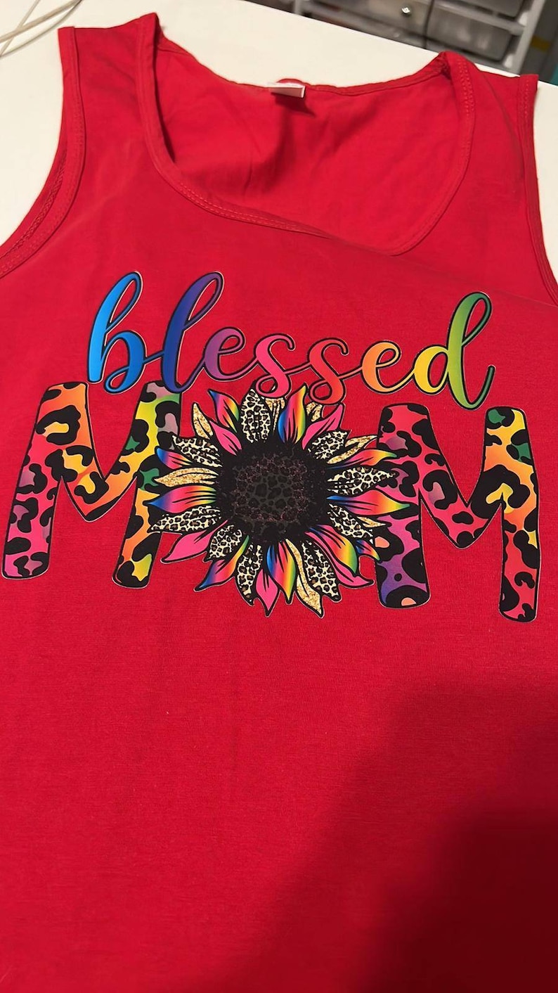 Puede incluir: Camiseta de tirantes roja con las palabras "Blessed Mom" en colores arco&iacute;ris con un girasol en el medio. El girasol tiene un centro negro y p&eacute;talos arco&iacute;ris con un estampado de leopardo.