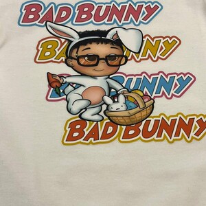 Puede incluir: Camiseta blanca con una imagen de dibujos animados de Bad Bunny vestido como un conejito de Pascua sosteniendo una cesta de huevos. El texto "Bad Bunny" se repite cuatro veces en diferentes colores alrededor de la imagen.