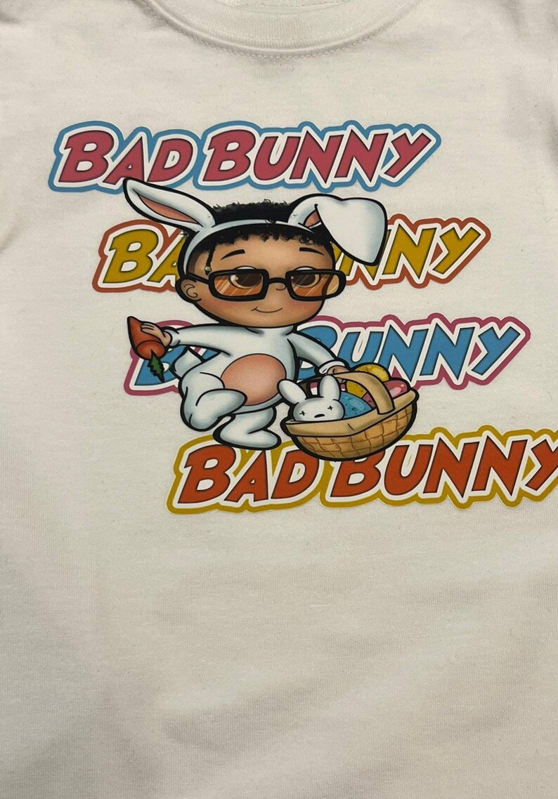 Puede incluir: Camiseta blanca con una imagen de dibujos animados de Bad Bunny vestido como un conejito de Pascua sosteniendo una cesta de huevos. El texto "Bad Bunny" se repite cuatro veces en diferentes colores alrededor de la imagen.