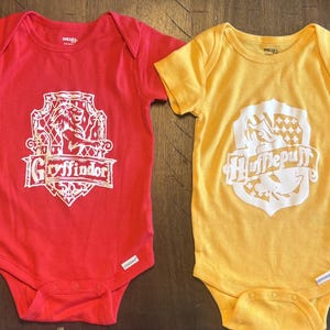 Puede incluir: Cuatro bodies de manga corta para bebé en verde, rojo, amarillo y azul. Cada uno presenta un escudo serigrafiado en blanco que representa las casas de Hogwarts: Slytherin con una serpiente, Gryffindor con un león, Hufflepuff con un tejón y Ravenclaw con un águila.