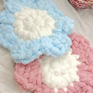 May include: Two chunky knit flower-shaped coasters or mats. One mat features light blue petals with a white center, and the other mat features dusty pink petals with a white center. Both items are crafted from thick, soft, plush yarn material.