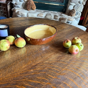 May include: An oval ceramic baking dish with a glossy brown exterior and a speckled light yellow interior. The dish is positioned on a dark wood table alongside a black candle jar labeled "OLET ENDS" and several small, round, green and red apples.