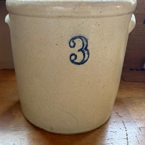 May include: A cylindrical stoneware crock with a beige, speckled finish and a thick, rounded rim. The front features a prominent, hand-painted blue numeral 3. Two small, integrated lug handles are positioned on the upper sides of the vessel.