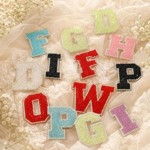 May include: A collection of varsity-style letter patches in various colors including light blue, lime green, pink, black, red, silver, and purple. Each letter is covered in sparkling rhinestones and bordered by a row of small clear crystals.