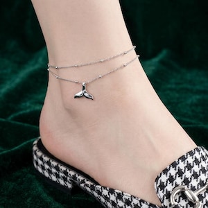 May include: A double-layered silver-toned anklet featuring a delicate chain with small evenly spaced beads and a central whale tail charm. The jewelry piece rests on the ankle, showcasing a polished metallic finish and a minimalist design.