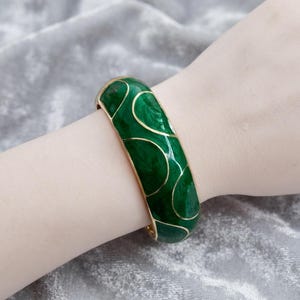 Könnte beinhalten: Ein breiter Armreif mit einer tief smaragdgrünen Emaille-Oberfläche und einer wirbelnden, marmorierten Textur. Der Armreif ist mit dünnen, polierten goldfarbenen Metalllinien akzentuiert, die organische, abgerundete Formen auf der gesamten Oberfläche bilden.
