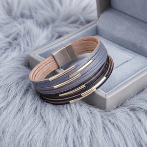 May include: A multi-strand bracelet featuring layered leather cords in shades of grey and black. The design includes polished gold-tone metal accents and a rectangular magnetic clasp closure, creating a structured and layered appearance.